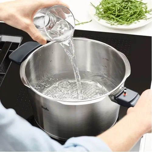 Stainless steel pot on a stove with water being poured into it, with a blurred background of a kitchen setting.