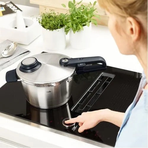 Stainless steel pressure cooker on a stovetop with a person interacting with it, in a kitchen setting.