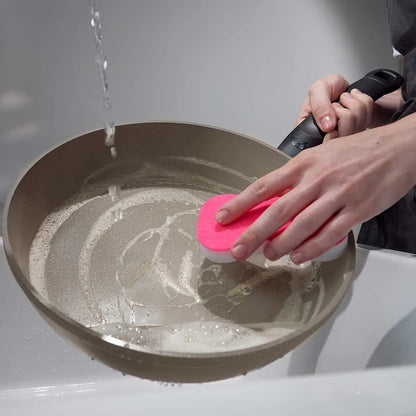 Person cleaning a frying pan with a pink scrubber under running water.