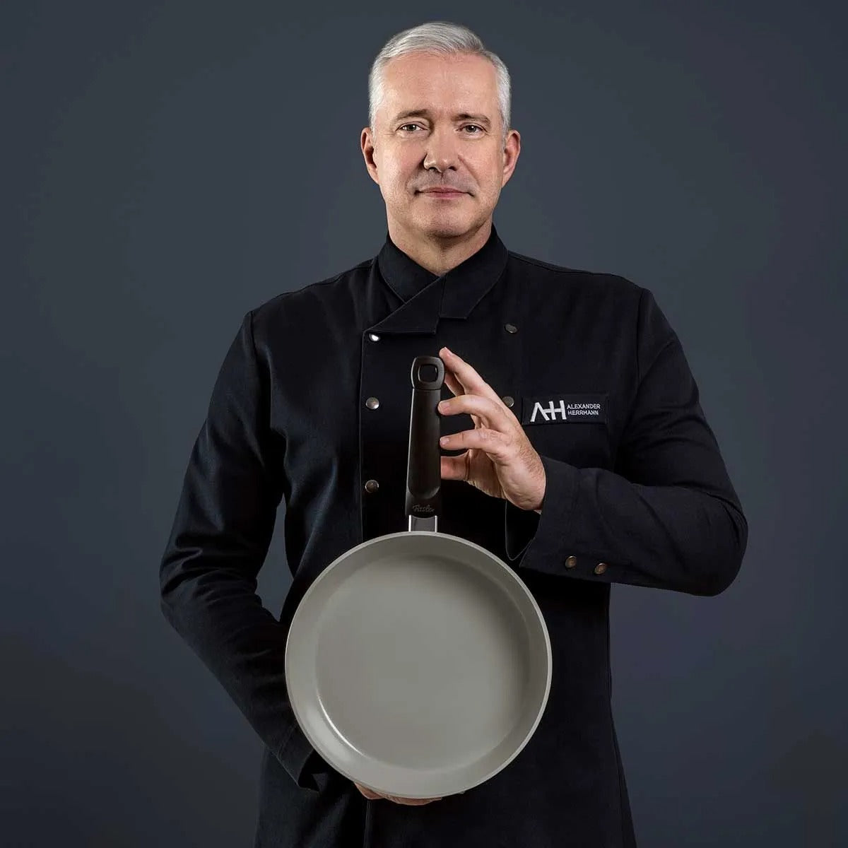 Man in a black chef's coat holding a gray frying pan against a dark background