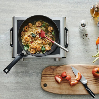 Frying pan with shrimp and pasta on a portable stove, surrounded by ingredients and utensils on a gray surface.