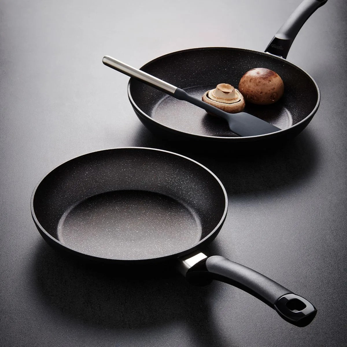 Two black frying pans on a gray surface with one containing food.